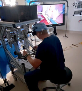 Dr. Ryan Broderick stă la o consolă de teleoperare și controlează un robot în timpul unei proceduri de extirpare a vezicii biliare.